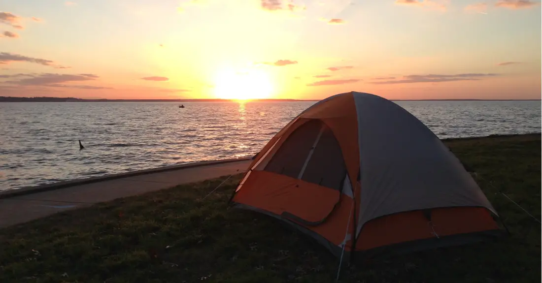 An orange tent pitched along the shore of Lake Livingston in Texas, with the sun setting over the lake in the background.